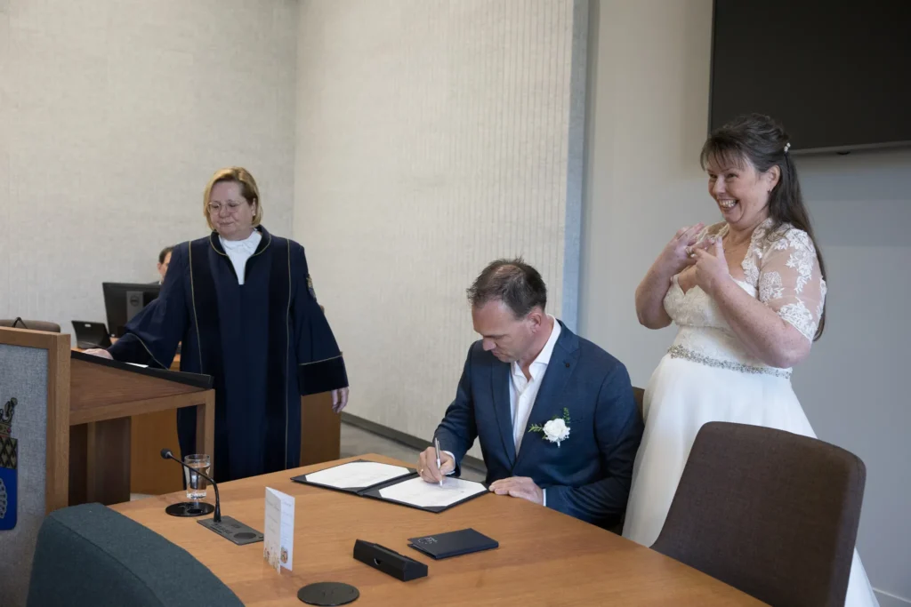 De bruidegom ondertekent de huwelijksakte tijdens de ceremonie terwijl de bruid er trots naast staat en enthousiast haar ring laat zien.